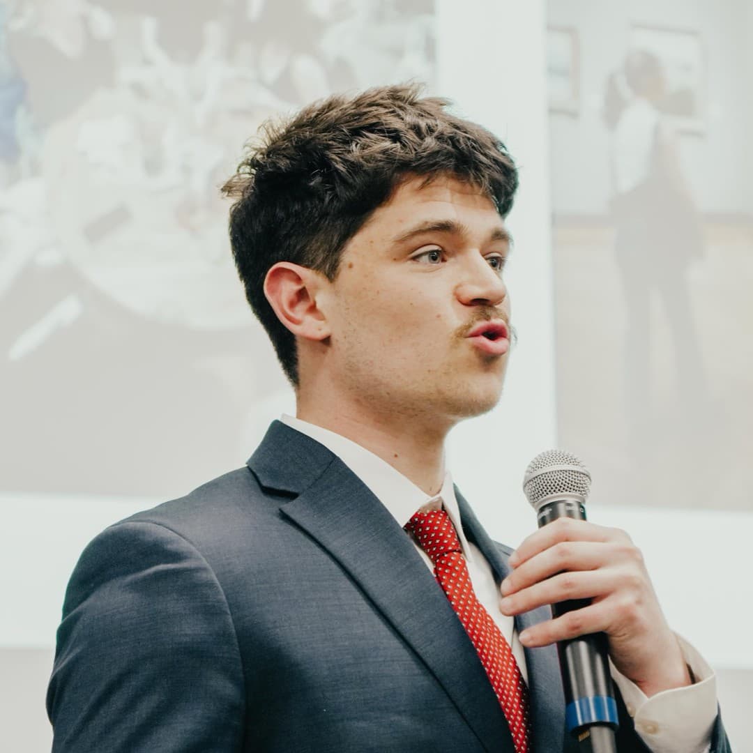 Judson Dunne in a navy suit and red tie, speaking into a microphone